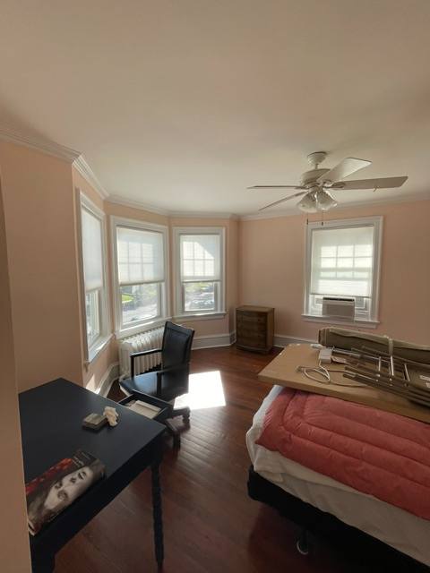 Bedroom with pink walls, wooden desk with office chair, bed with red comforter, ceiling fan, and bay windows